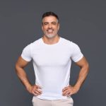 Happy confident handsome fit sporty healthy mature older man, middle aged sportsman athlete or personal trainer wearing white t-shirt standing isolated on gray background advertising gym. Portrait